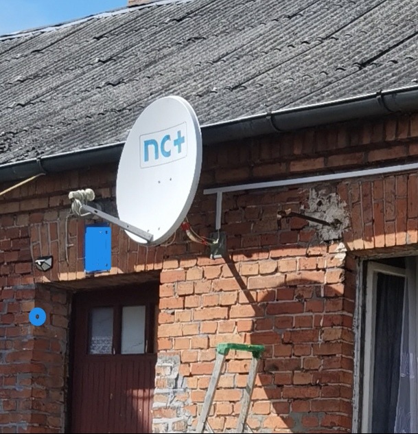 Biała antena satelitarna z logo nc+ zamontowana na ceglanej ścianie budynku, widoczna drabina oparta o ścianę, szary dach z rynnami w tle.