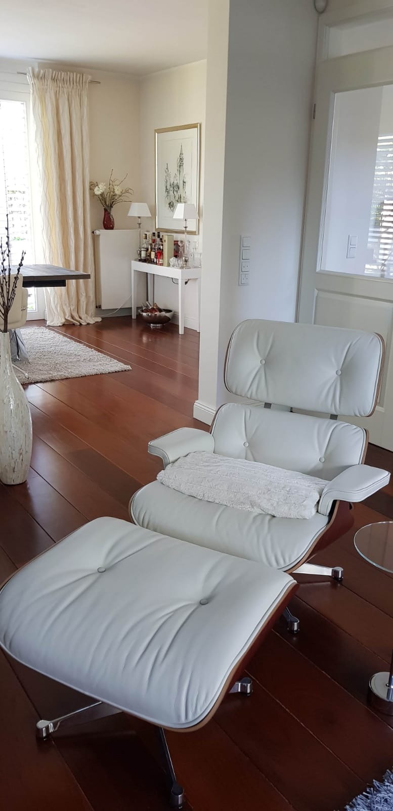 Biały, skórzany fotel z podnóżkiem w nowoczesnym salonie z drewnianą podłogą i jasnymi ścianami. W tle widoczny stół z bukietem w wazonie, biały kredens z alkoholem i dekoracjami oraz okno...