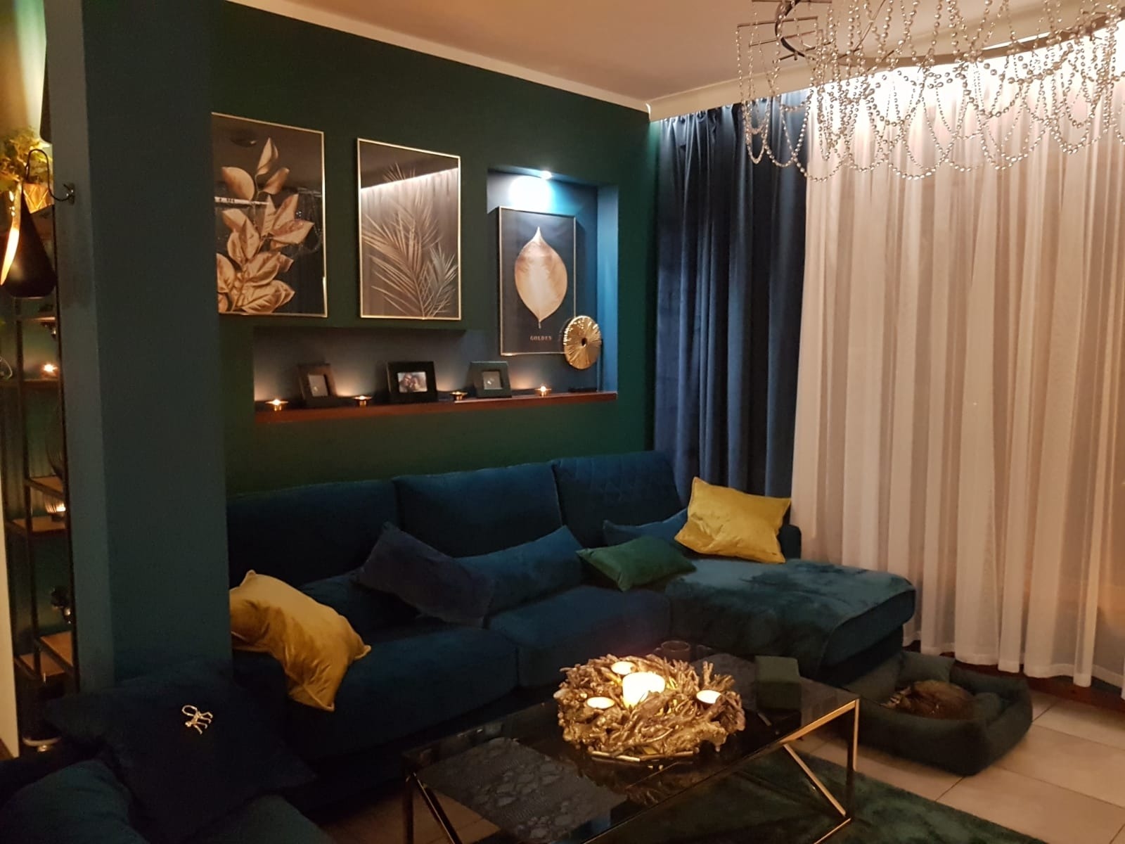 Salon w odcieniach butelkowej zieleni i granatu z welurową sofą, szklanym stolikiem kawowym ze złotym stelażem, dekoracyjnym wieńcem ze świecami i kryształowym żyrandolem.