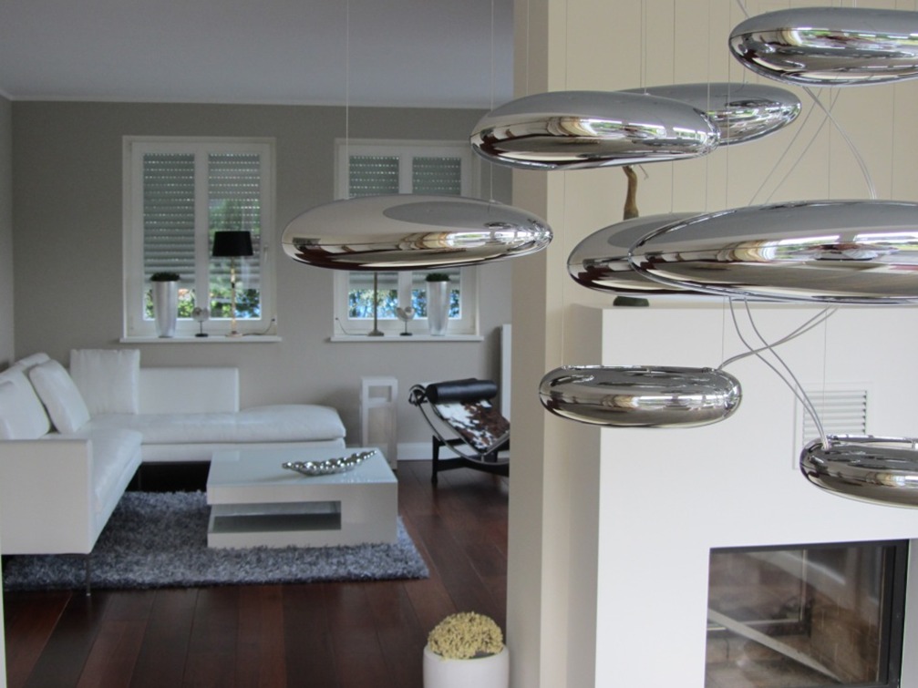 Nowoczesny salon z białym narożnikiem, szarym dywanem i podłogą z ciemnego drewna, oświetlony designerskimi lampami w kształcie srebrnych dysków, z kominkiem w tle.