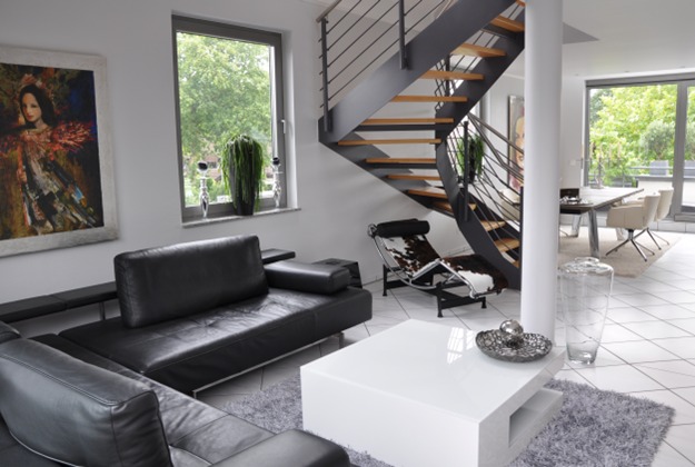 Jasne, nowoczesne wnętrze domu z czarną skórzaną sofą, białym stolikiem kawowym, spiralnymi schodami z drewnianymi stopniami i metalowymi poręczami, obrazem na ścianie i widokiem na zieleń za oknem.