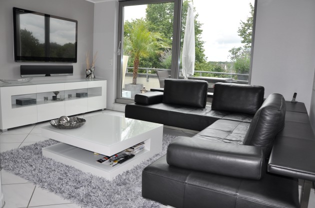 Nowoczesny salon w szarościach: czarna skórzana sofa narożna, biały stolik kawowy z gazetami na półce, biała szafka RTV z dekoracjami i telewizorem, widok na zielony balkon z palmą.