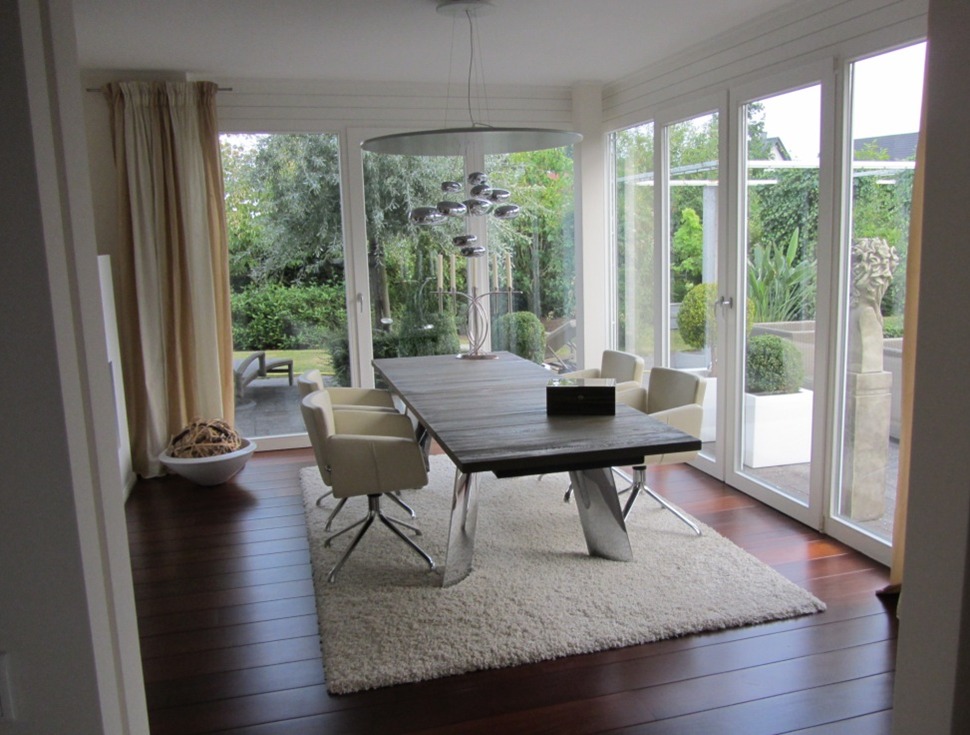 Nowoczesna jadalnia z dużym stołem z ciemnego drewna, krzesłami z jasnej skóry na metalowych nogach i puszystym dywanem, widok na zadbany ogród przez panoramiczne okna.