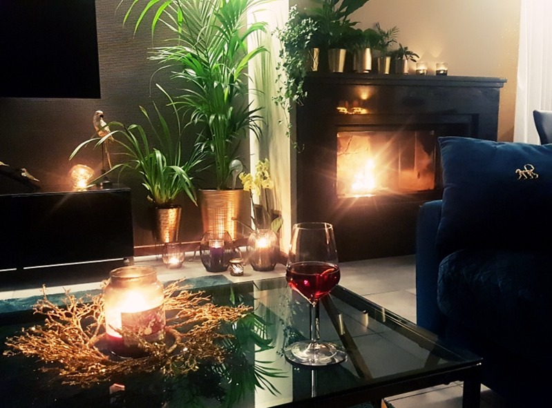Przytulne wnętrze salonu z kominkiem, roślinami w donicach, świecami i kieliszkiem czerwonego wina na szklanym stoliku kawowym, oświetlone ciepłym światłem.