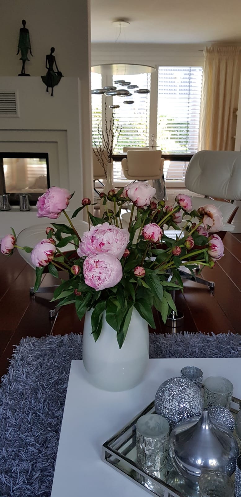 Bukiet różowych piwonii w białym wazonie na białym stoliku kawowym z szarym dywanem, w tle salon z kominkiem i nowoczesnymi meblami.