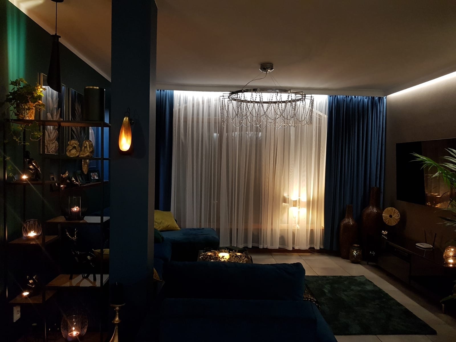 Ciemne, nastrojowe wnętrze salonu z granatowymi ścianami, oświetlone świecami i lampami, z regałem pełnym dekoracji, okrągłym żyrandolem z łańcuszków i turkusową sofą