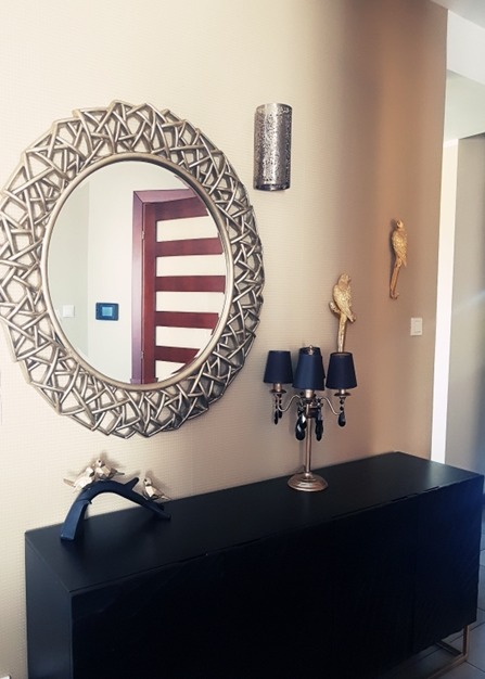 Czarna komoda z dekoracjami: okrągłe lustro w ozdobnej ramie, lampka z czarnymi abażurami i kryształkami, srebrne figurki ptaków i złote figurki papug na ścianie.