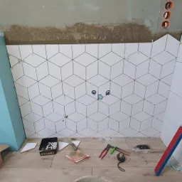 Ściana łazienki w trakcie układania białych płytek o geometrycznym wzorze, widoczne narzędzia glazurnicze i wiadro z zaprawą na podłodze imitującej drewno.