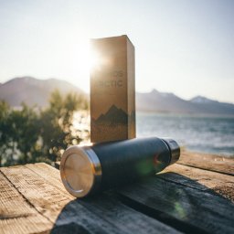 Piotr Widomski - Czarny termos i kartonowe pudełko na drewnianym stole z widokiem na morze i góry w tle, oświetlone promieniami słońca.