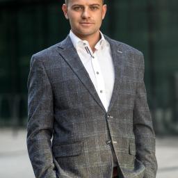 Piotr Widomski - Mężczyzna w szarej, kraciastej marynarce i białej koszuli, z rękami w kieszeniach spodni, pozuje na tle szklanej fasady budynku.