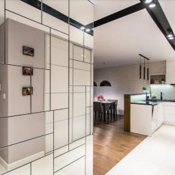 Piotr Widomski - Nowoczesne wnętrze: lustrzana ściana z geometrycznym podziałem, drewniane drzwi wejściowe, białe płytki podłogowe, widok na jadalnię i kuchnię z białymi meblami.