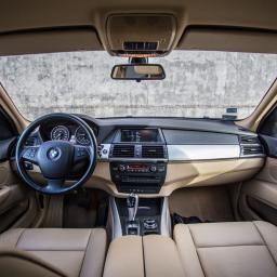 Piotr Widomski - Wnętrze beżowego BMW X5 z widokiem na deskę rozdzielczą, kierownicę z logo i skórzane fotele, na tle betonowej ściany.