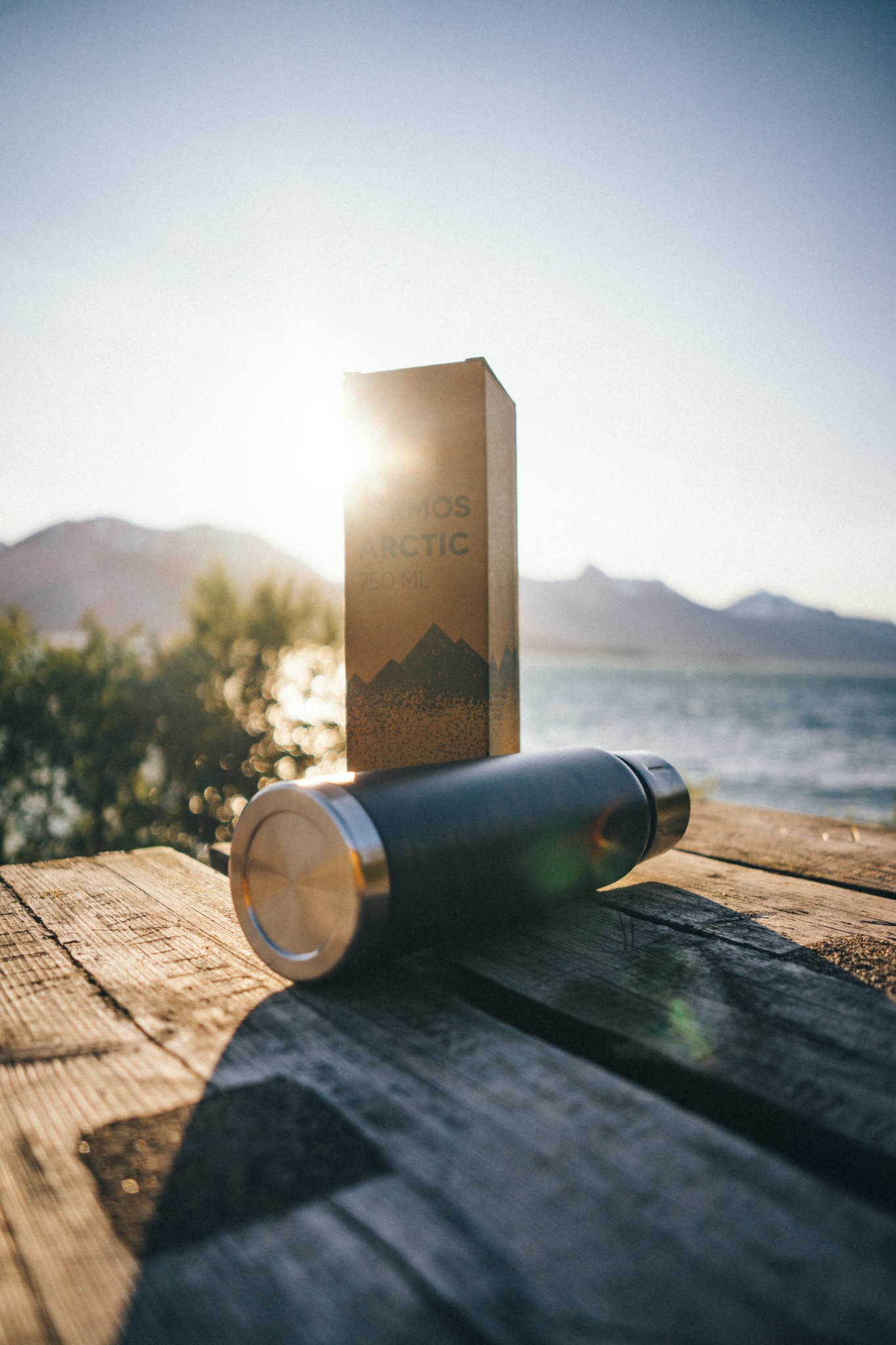 Czarny termos i kartonowe pudełko na drewnianym stole z widokiem na morze i góry w tle, oświetlone promieniami słońca.