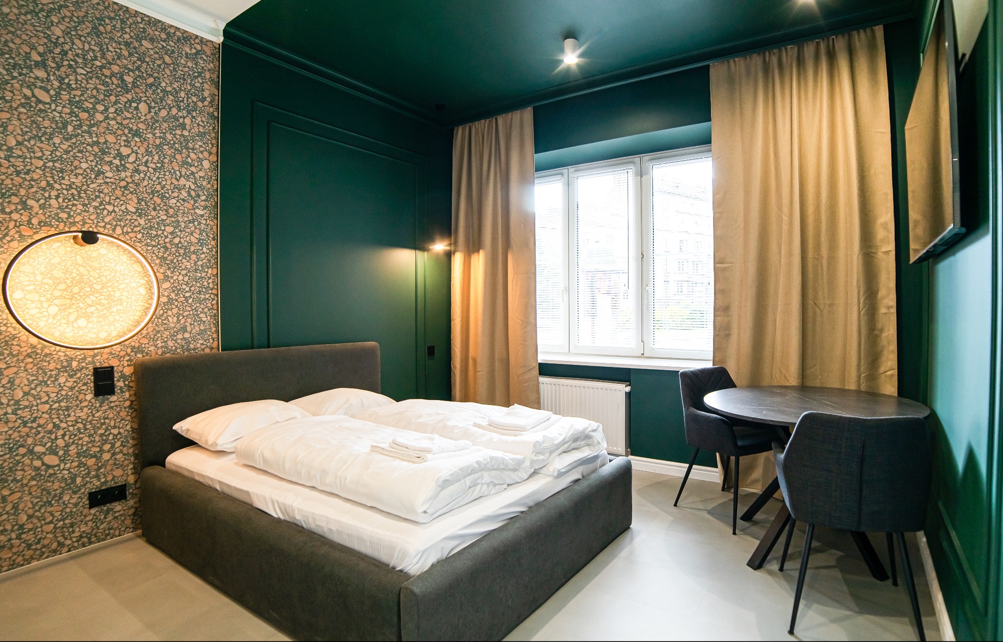 Pokój hotelowy z ciemnozielonymi ścianami, łóżkiem z szarą ramą i białą pościelą, okrągłą lampą na ścianie o wzorzystej tapecie, stolikiem z krzesłami pod oknem i zasłonami.