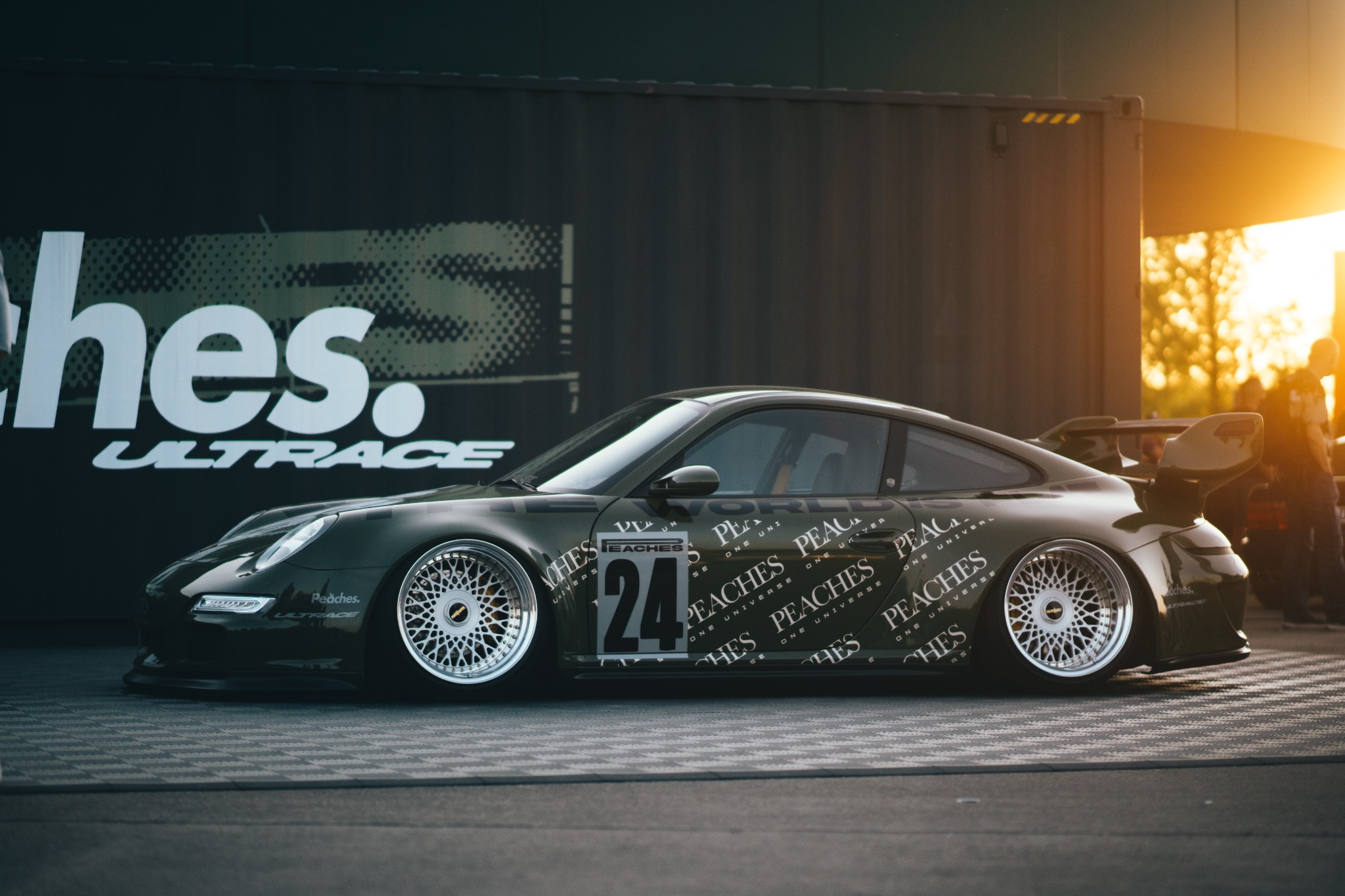 Olive green Porsche 911 with custom wheels, lowered suspension, and 'Peaches' livery parked in front of a container with 'Ultrace' branding, bathed in golden hour sunlight.