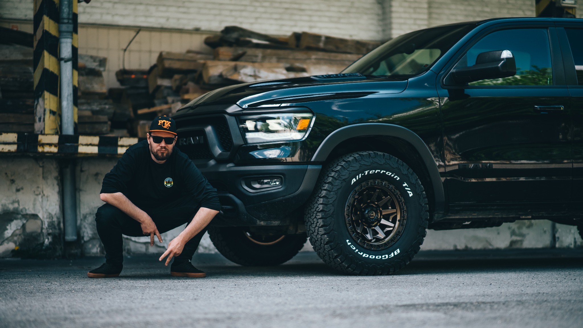 Mężczyzna w czarnym ubraniu kuca obok czarnego samochodu RAM z oponami terenowymi, w tle paleta desek i ściana z żółto-czarnymi pasami.