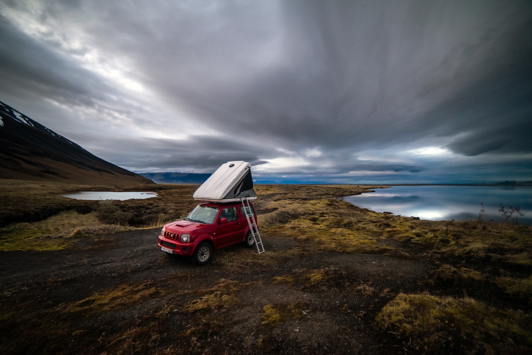 Czerwony samochód terenowy Suzuki Jimny z rozkładanym białym namiotem dachowym i drabinką na tle surowego islandzkiego krajobrazu z jeziorem i pochmurnym niebem.