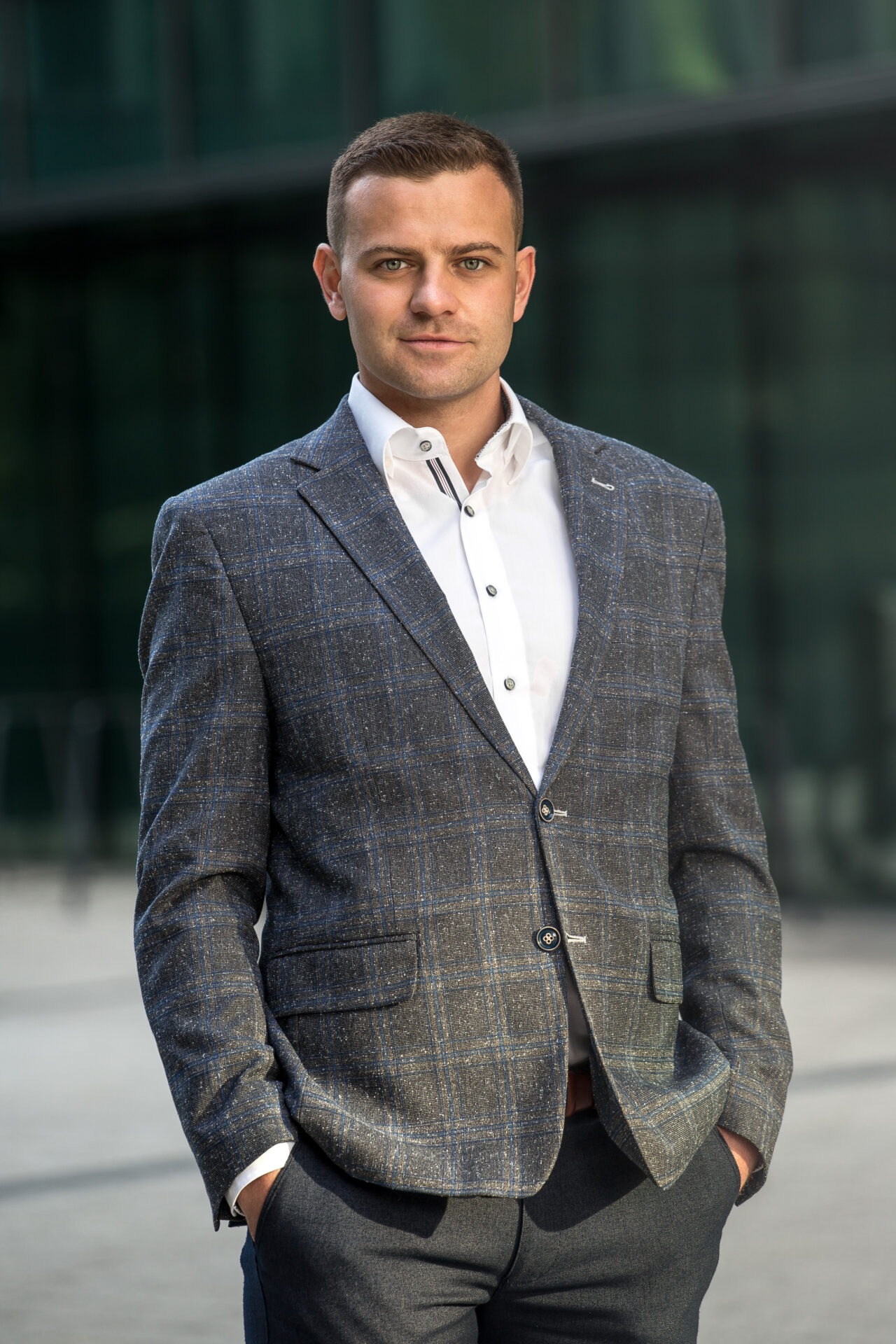 Mężczyzna w szarej, kraciastej marynarce i białej koszuli, z rękami w kieszeniach spodni, pozuje na tle szklanej fasady budynku.