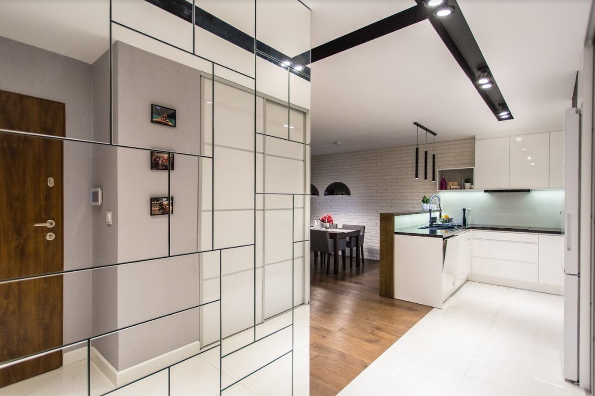 Nowoczesne wnętrze: lustrzana ściana z geometrycznym podziałem, drewniane drzwi wejściowe, białe płytki podłogowe, widok na jadalnię i kuchnię z białymi meblami.