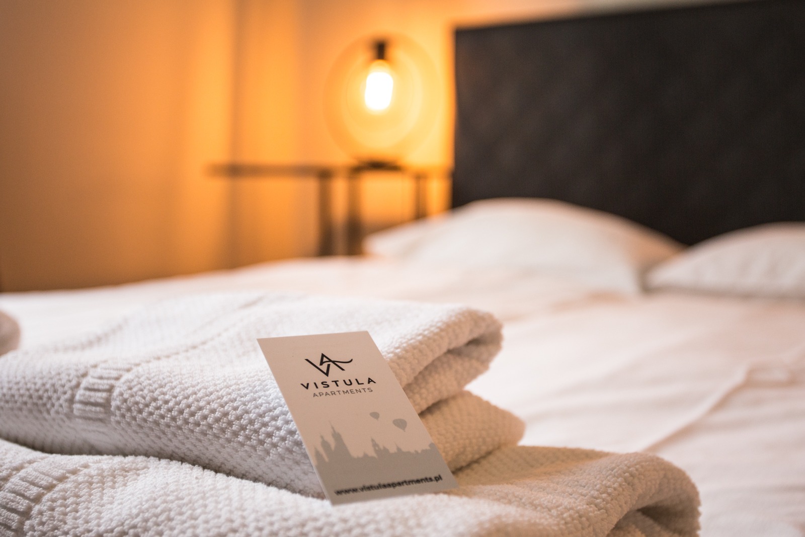 Stos białych, puszystych ręczników z wizytówką Vistula Apartments na łóżku w pokoju hotelowym z oświetleniem w tle.
