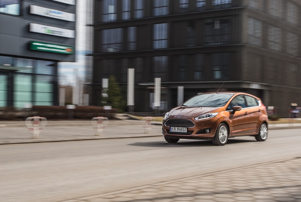 Brązowy Ford Fiesta w ruchu na tle nowoczesnego budynku biurowego z szarymi panelami.