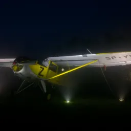 Żółto-srebrny samolot SP-YWZ oświetlony reflektorami na tle nocnego nieba i lasu.
