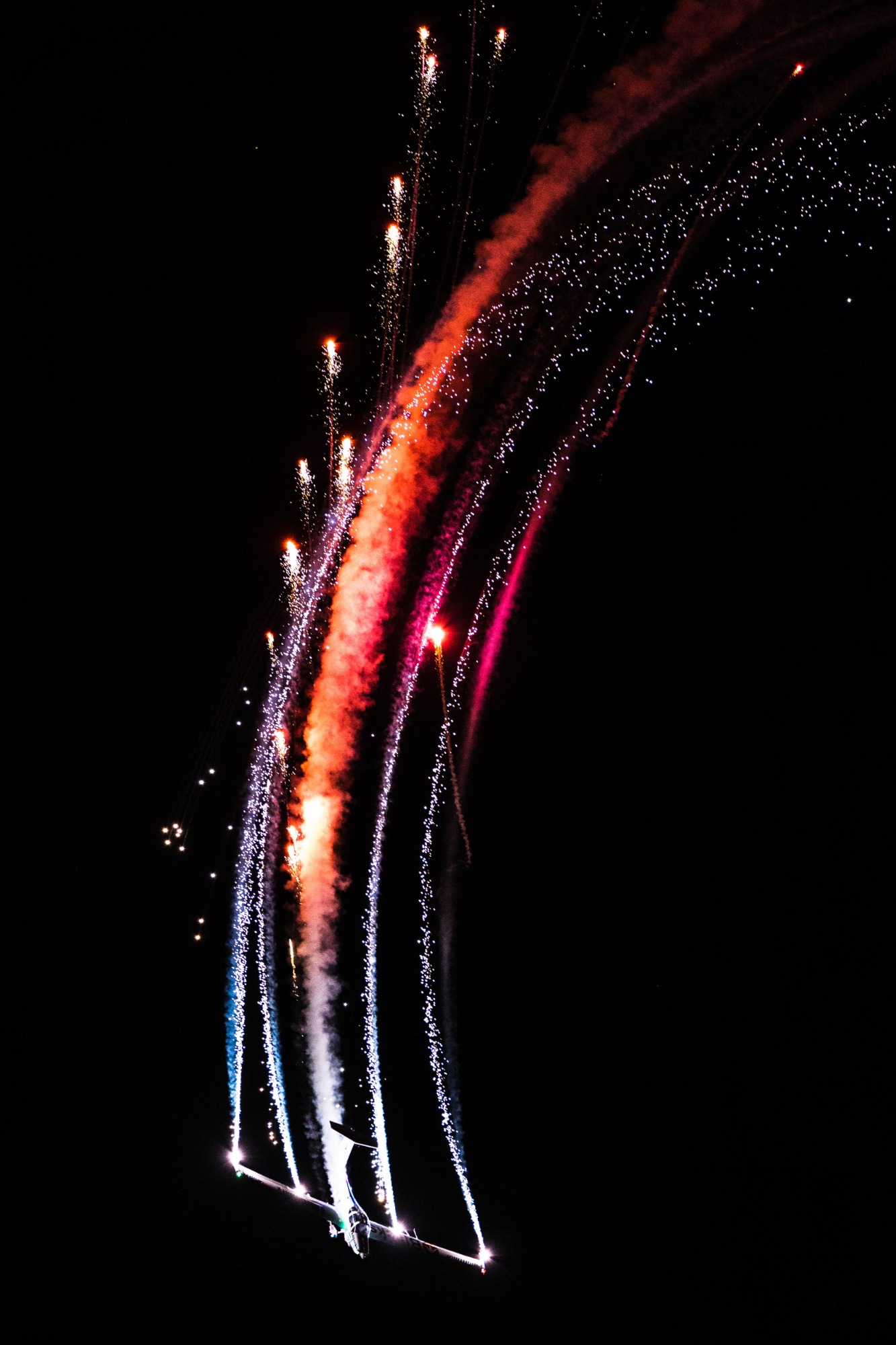 Akrobacje samolotu w Bydgoszczy z pirotechniką, tworzące barwne smugi na nocnym niebie.
