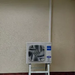 Zewnętrzna jednostka klimatyzacji Haier na białym metalowym stojaku, przymocowana do beżowej ściany budynku z ceglaną podmurówką, z widoczną białą rurą odprowadzającą, na tle zieleni.