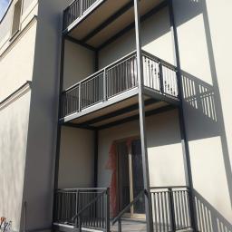 GOWIMET - Trzypoziomowe balkony z szarą metalową balustradą na tle jasnej elewacji budynku w trakcie budowy, widoczne narzędzia budowlane przy ścianie.