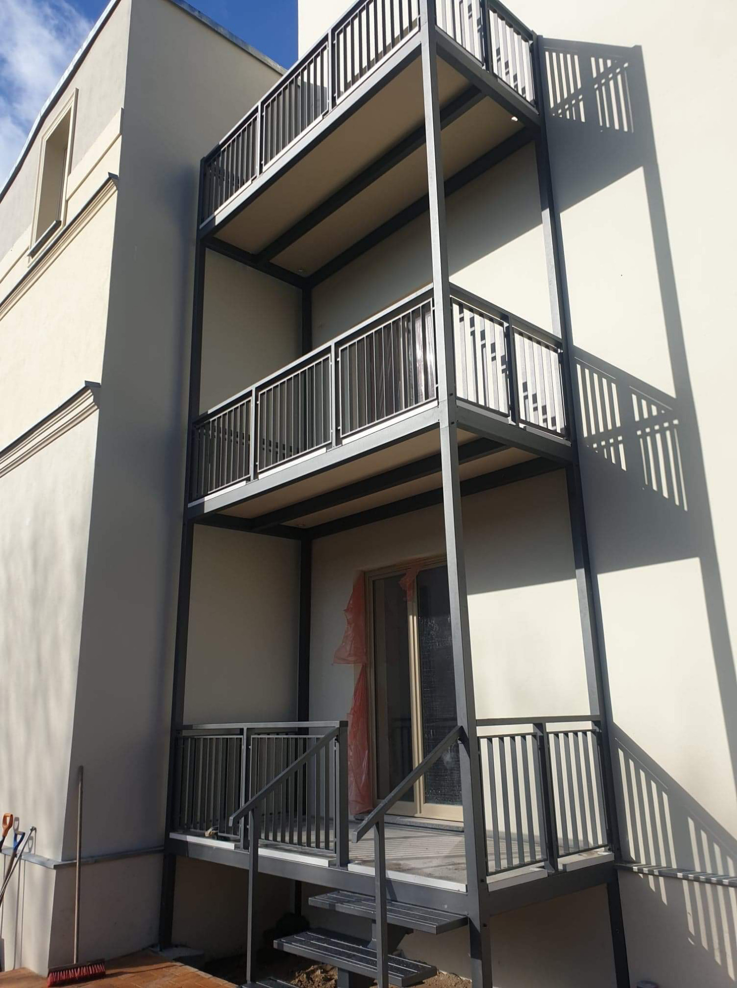 Trzypoziomowe balkony z szarą metalową balustradą na tle jasnej elewacji budynku w trakcie budowy, widoczne narzędzia budowlane przy ścianie.