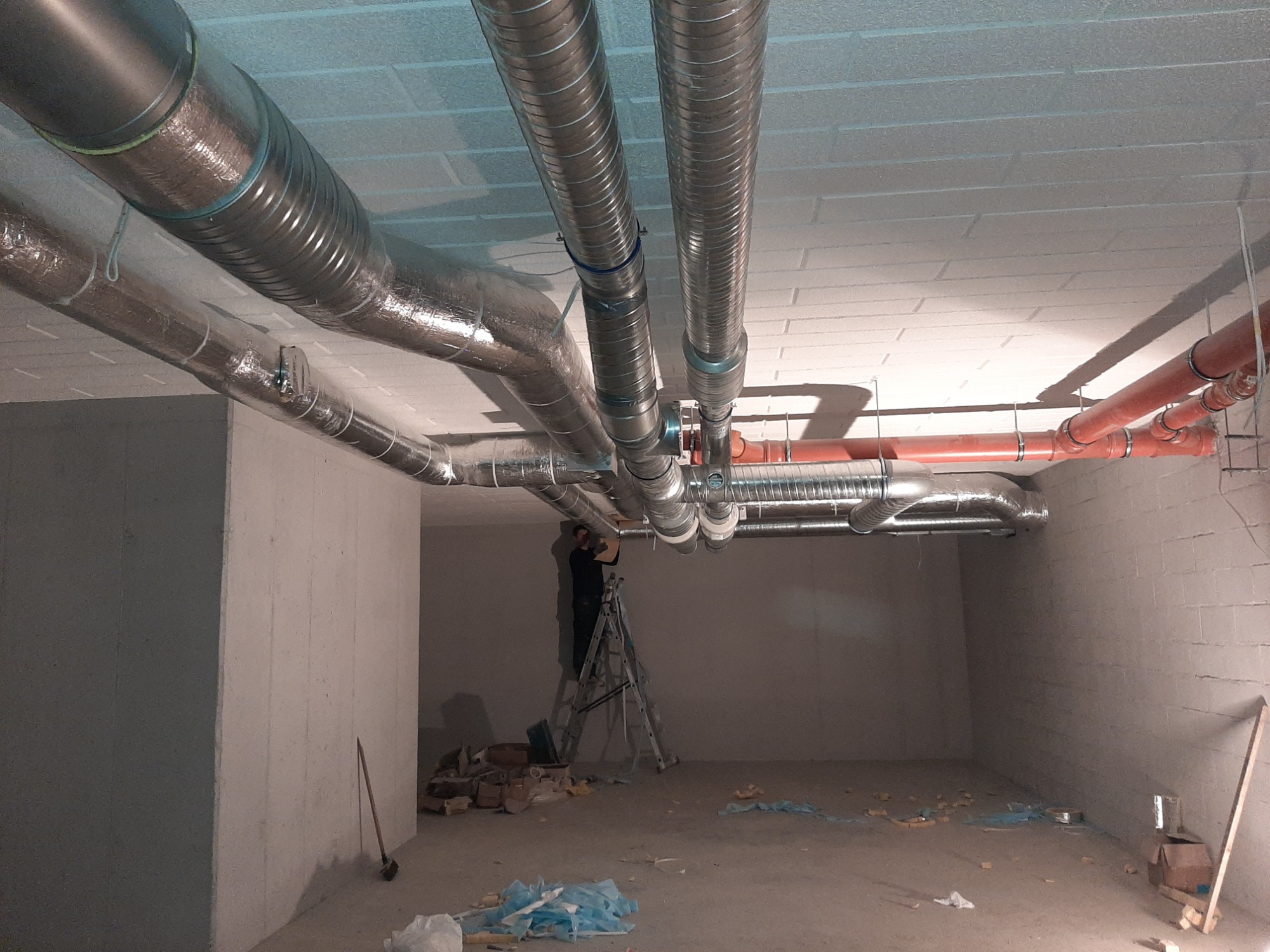 Instalacja wentylacji: widok na sufit z zamontowanymi, metalowymi rurami wentylacyjnymi, część z izolacją, pracownik na drabinie montuje system, widoczne narzędzia i ślady po pracy.