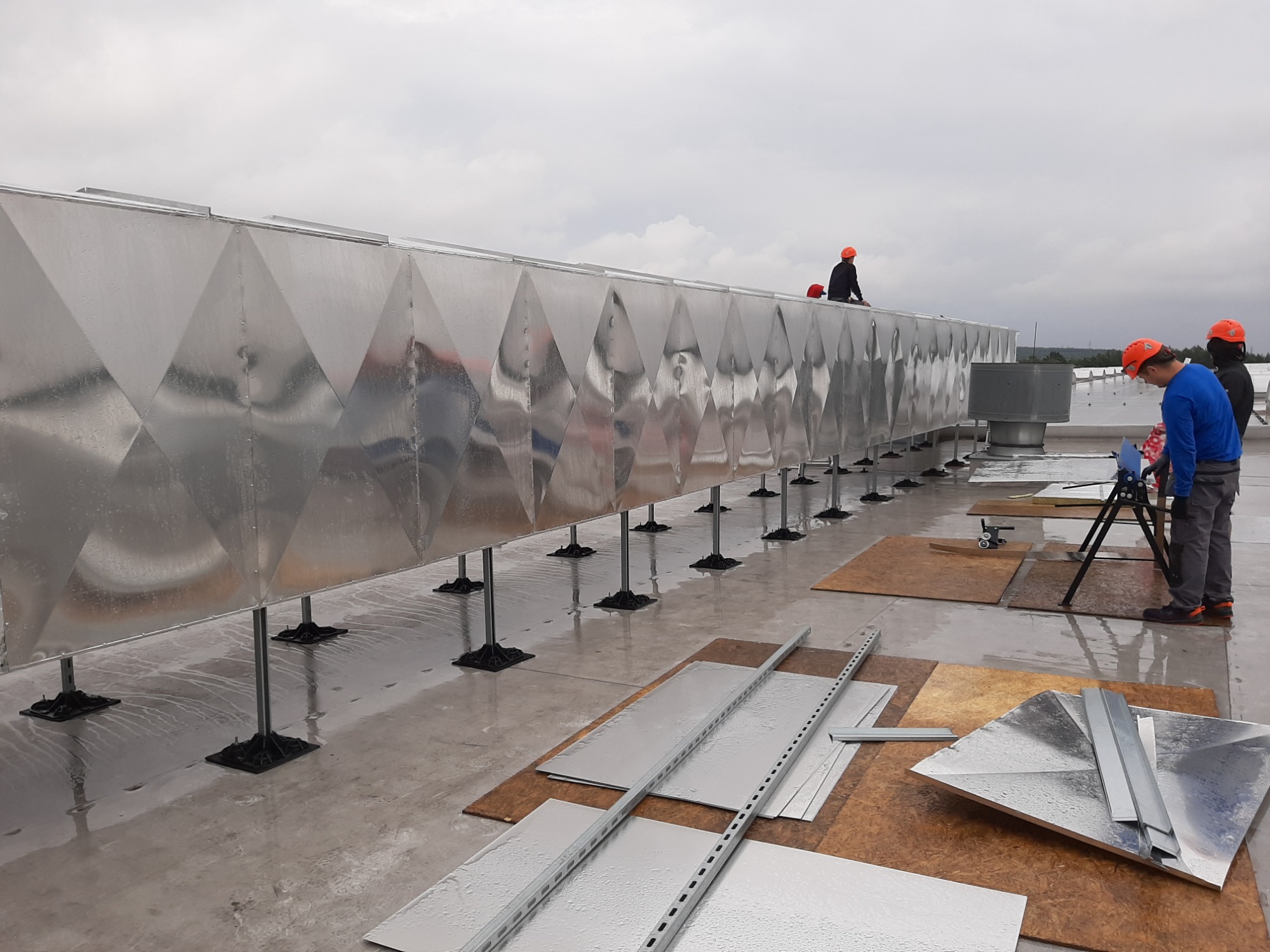 Długa, srebrna instalacja wentylacyjna z romboidalnym wzorem, umieszczona na dachu budynku, wsparta na czarnych podstawach. Trzech pracowników w pomarańczowych kaskach pracuje przy instalacji...