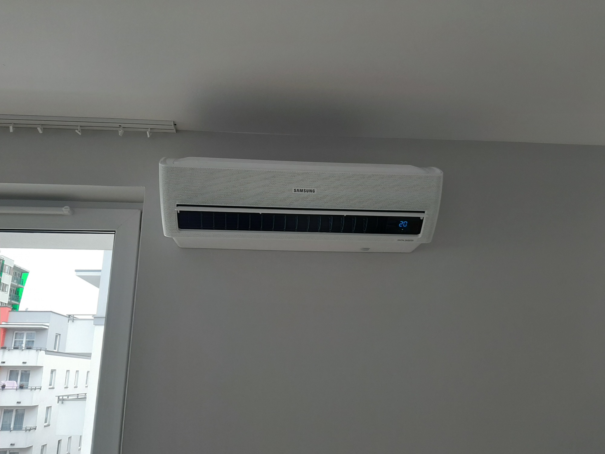 Biała klimatyzacja Samsung zamontowana na szarej ścianie w mieszkaniu, widok z boku okna z widokiem na budynek mieszkalny.