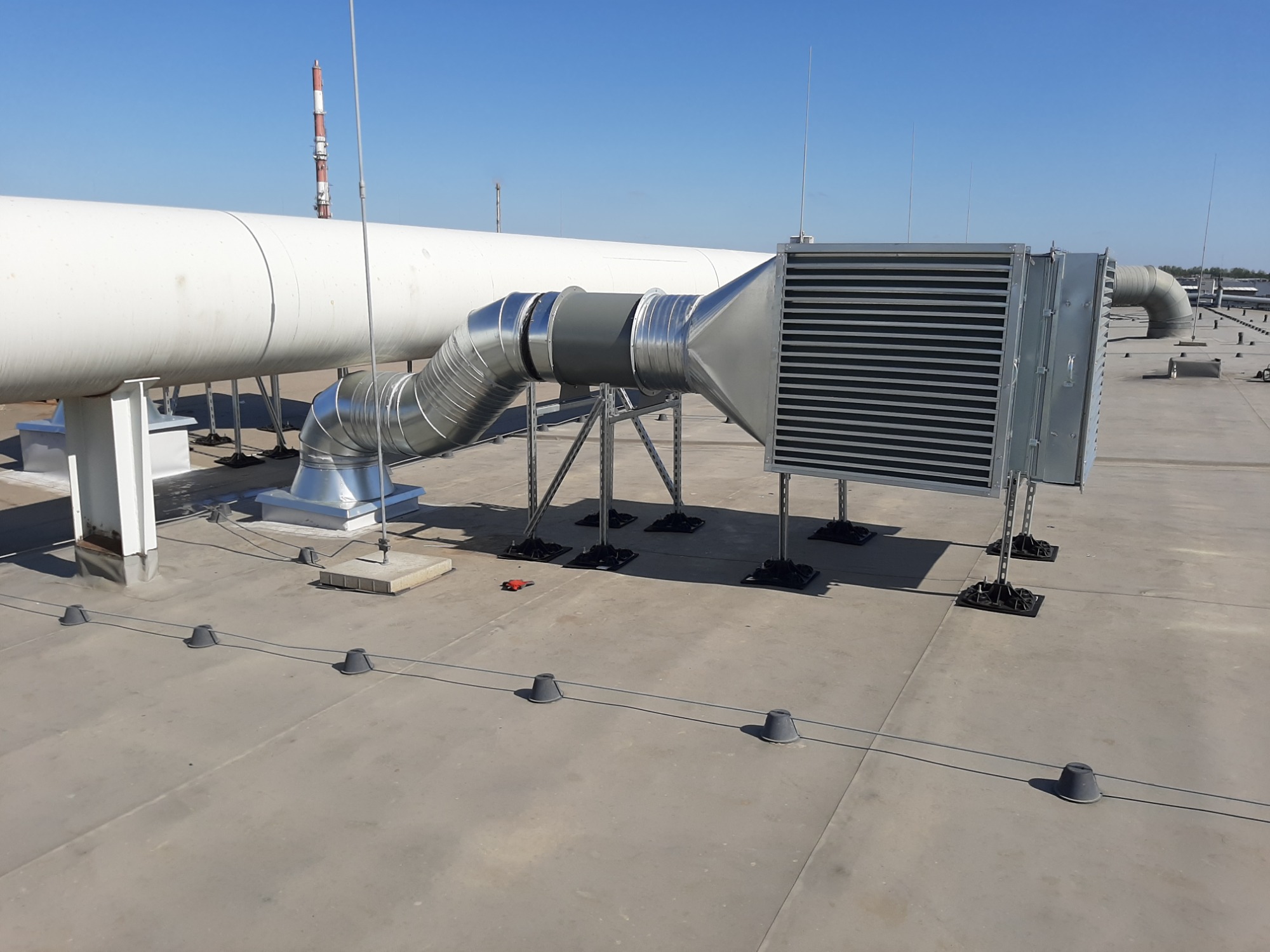 System wentylacyjny z metalowymi rurami, filtrem i wspornikami na płaskim dachu budynku przemysłowego, widoczny przewód odgromowy i komin w tle.