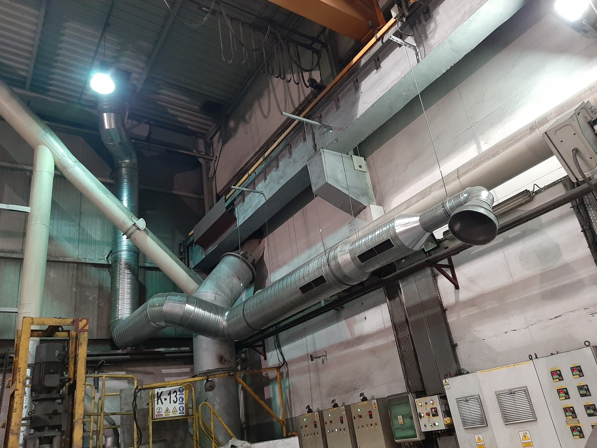 Kompleksowy system wentylacji z metalowymi rurami i kanałami w przestrzeni przemysłowej, widoczne elementy sterujące na ścianie oraz żółte barierki ochronne.