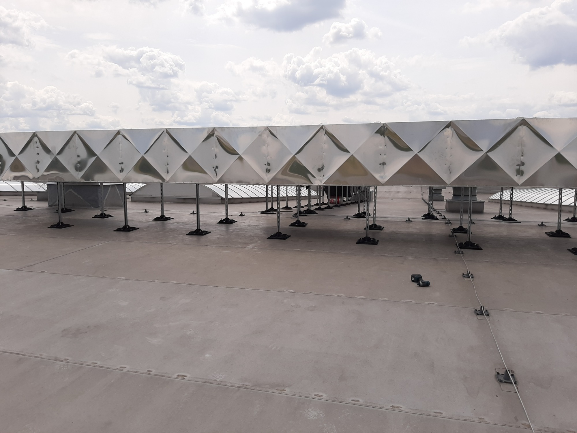 Błyszczący kanał wentylacyjny o diamentowym wzorze, umieszczony na dachu budynku komercyjnego, wsparty na metalowych nogach z czarnymi podstawami, pod pochmurnym niebem.