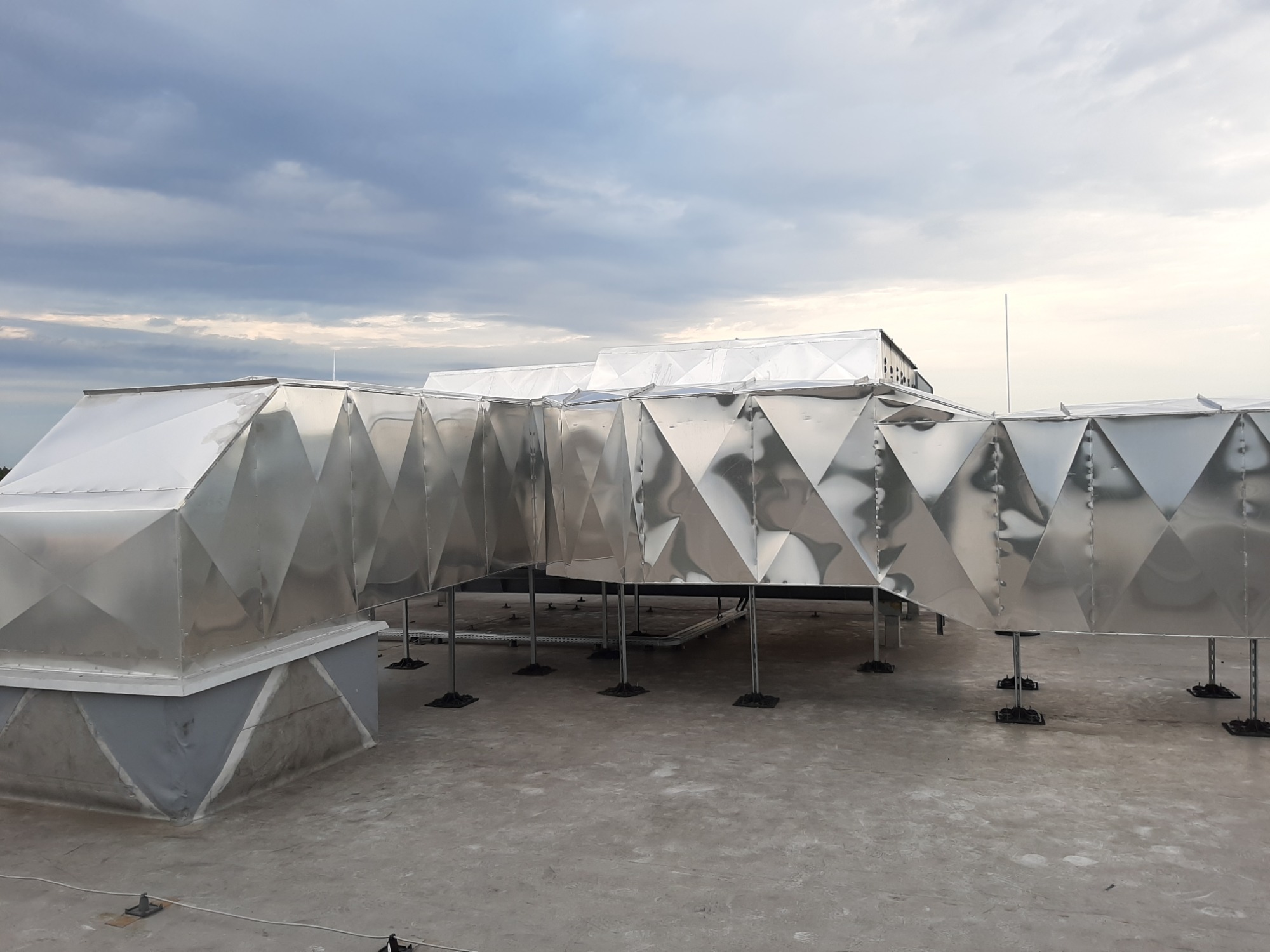 System kanałów wentylacyjnych z metalu na dachu budynku, widoczne podparcia i łączenia segmentów, pochmurne niebo w tle.