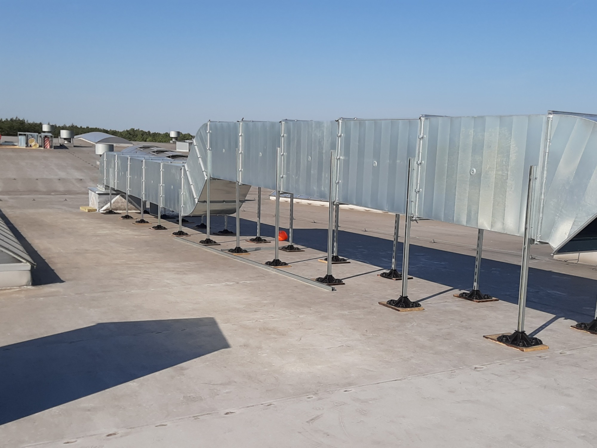 System kanałów wentylacyjnych z ocynkowanej blachy, zamontowany na dachu budynku komercyjnego na podporach z czarnego tworzywa, widok z góry w słoneczny dzień.