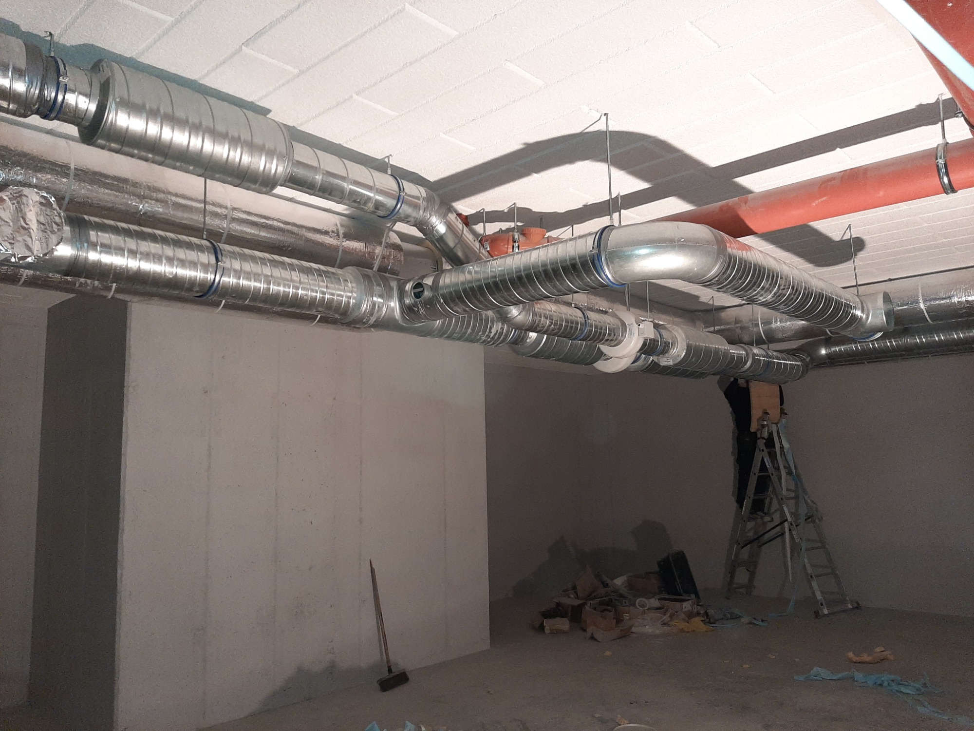 Instalacja spiralnych przewodów wentylacyjnych pod sufitem w surowym wnętrzu, widoczna drabina i postać w tle, elementy izolacji na przewodach.