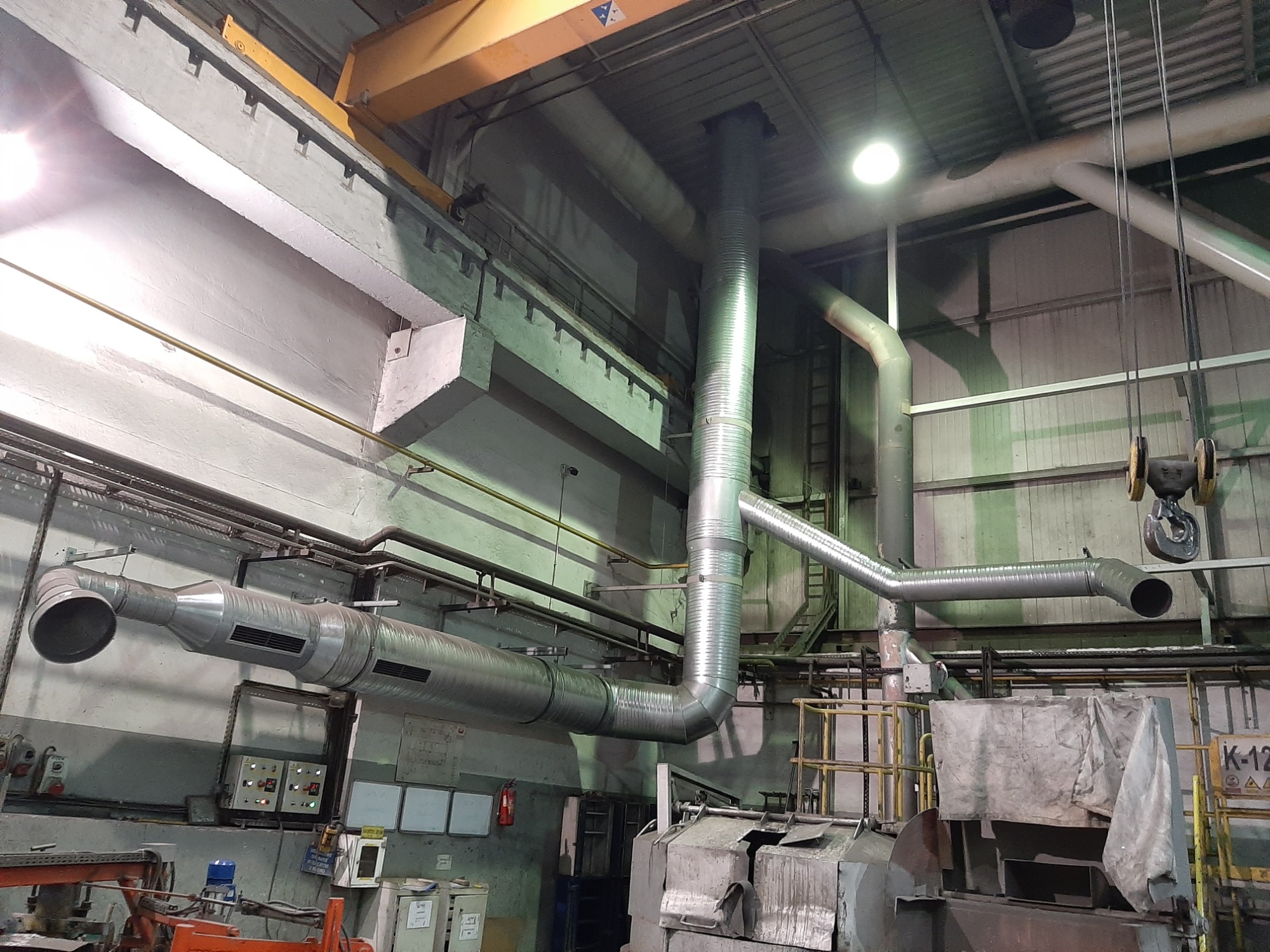 System wentylacji przemysłowej z metalowymi rurami, widoczne elementy konstrukcyjne hali, oświetlenie sufitowe i fragment maszyny osłoniętej materiałem.