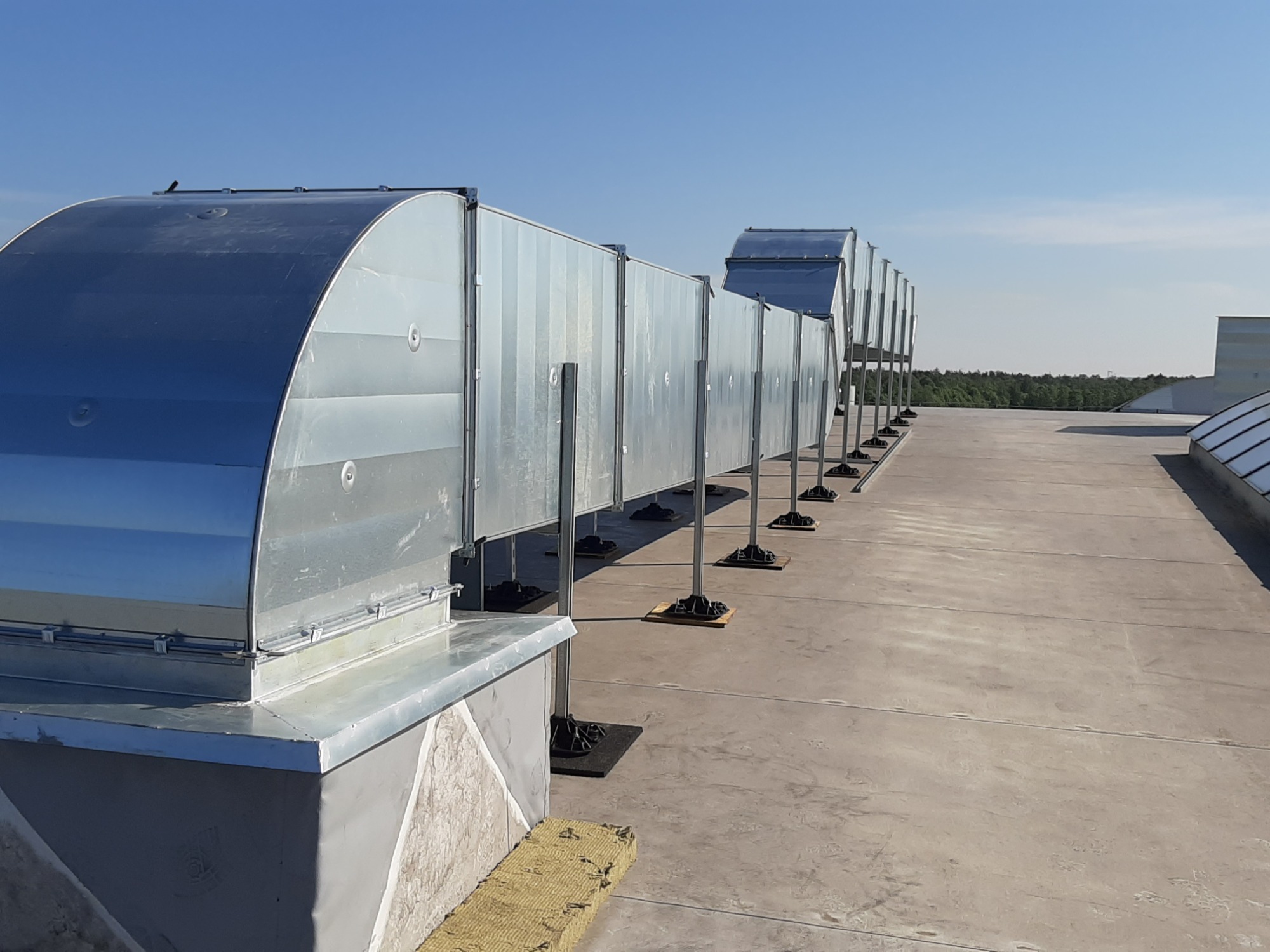 Długi ciąg srebrnych, prostokątnych kanałów wentylacyjnych na płaskim dachu budynku, z widoczną izolacją z wełny mineralnej u podstawy pierwszego elementu i lasem w tle pod błękitnym niebem.
