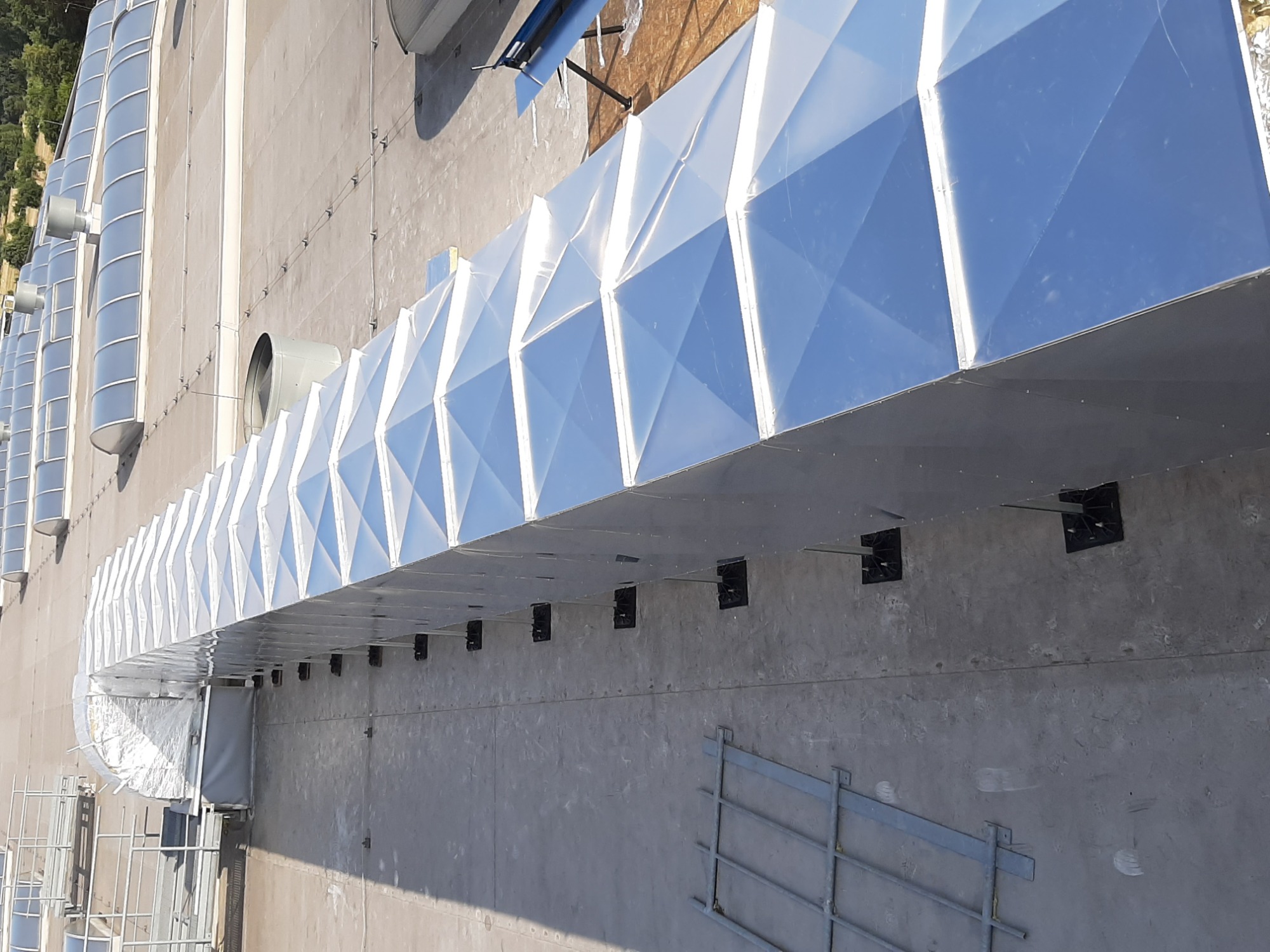 Zewnętrzny system kanałów wentylacyjnych o diamentowej strukturze, zamontowany na szarej ścianie budynku komercyjnego, z widocznymi wspornikami montażowymi i elementami konstrukcyjnymi dachu w tle,...