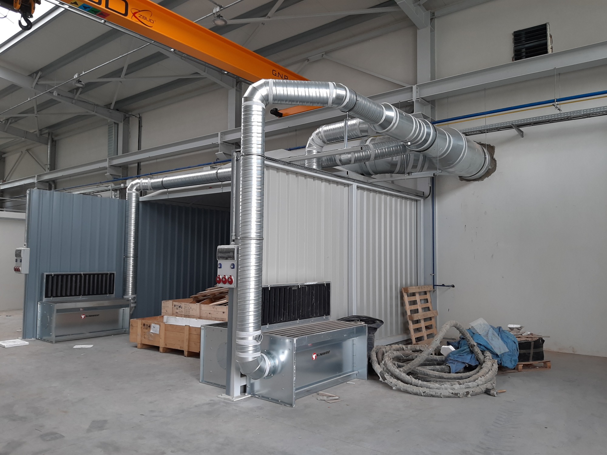 System kanałów wentylacyjnych z ocynkowanej blachy w hali przemysłowej, widoczny suwnica pomarańczowa pod sufitem, skrzynie drewniane i zwój kabli na podłodze.