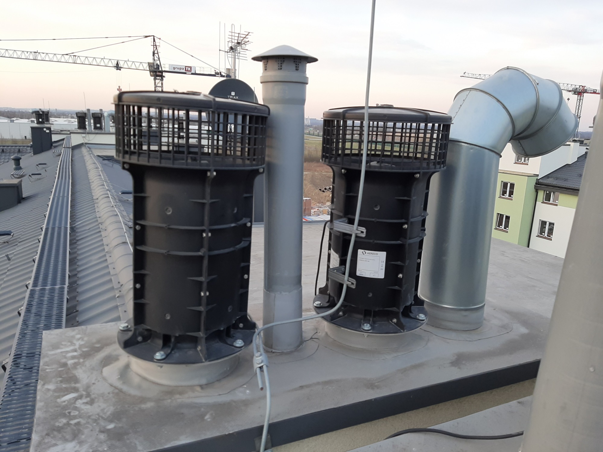 Dwa czarne wentylatory dachowe z metalowymi kominami na płaskim dachu w Nowej Dębie, z widocznymi budynkami i żurawiami w tle.