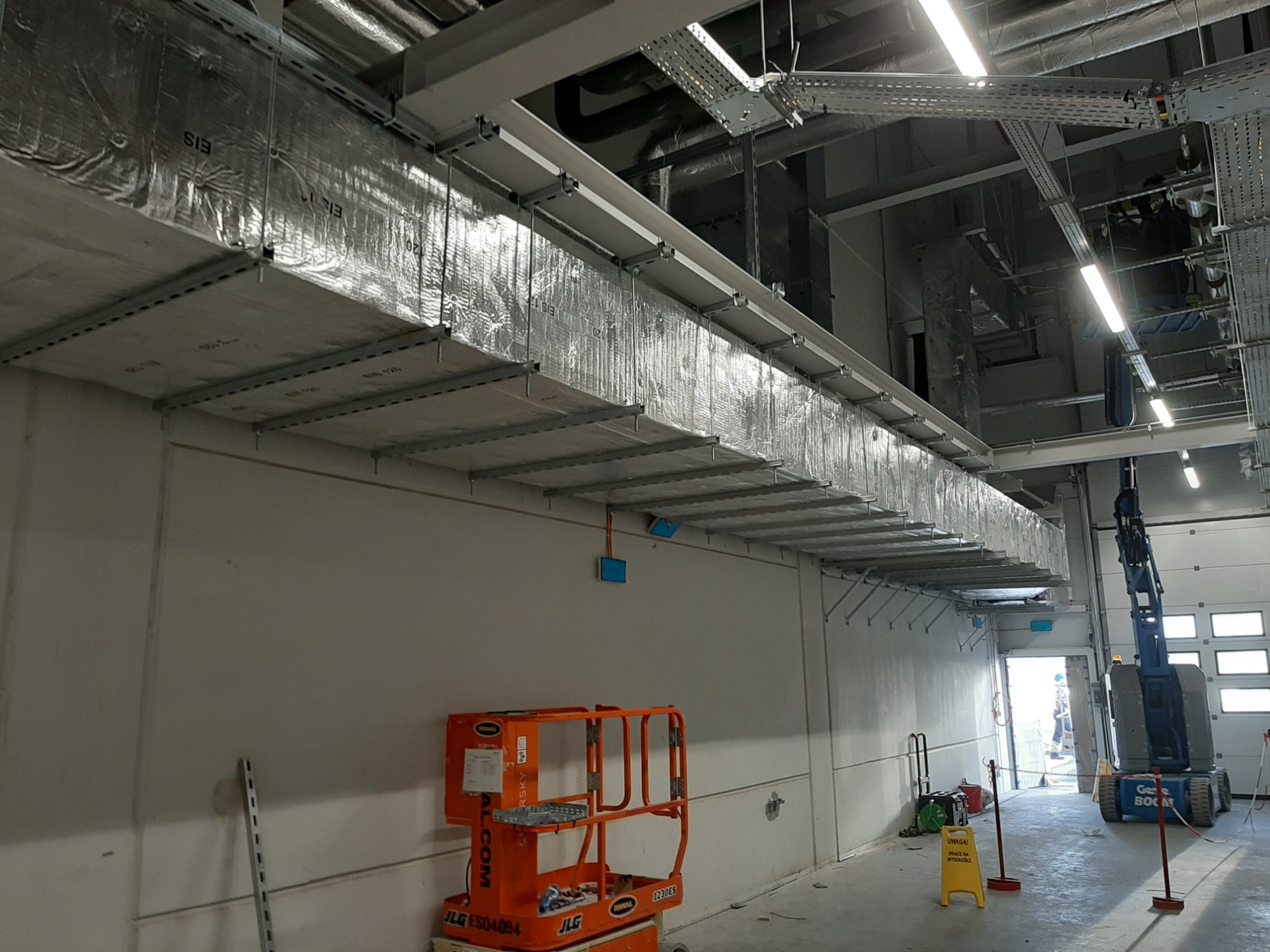 Srebrna, zaizolowana kanałowa instalacja wentylacyjna pod sufitem w budynku komercyjnym, widoczny podnośnik koszowy i tablica ostrzegawcza.