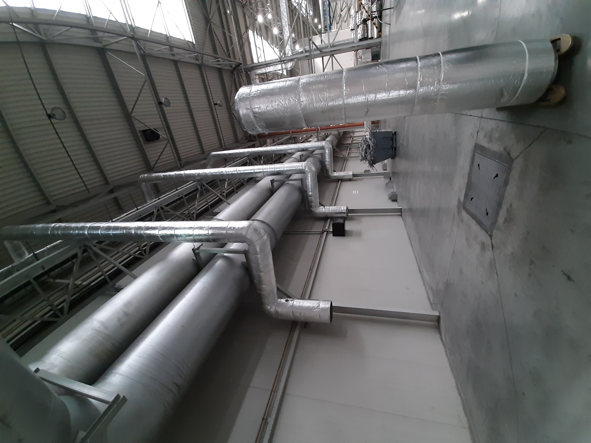 System rur wentylacyjnych z izolacją termiczną, zamontowany pod sufitem hali przemysłowej, widok z dołu.