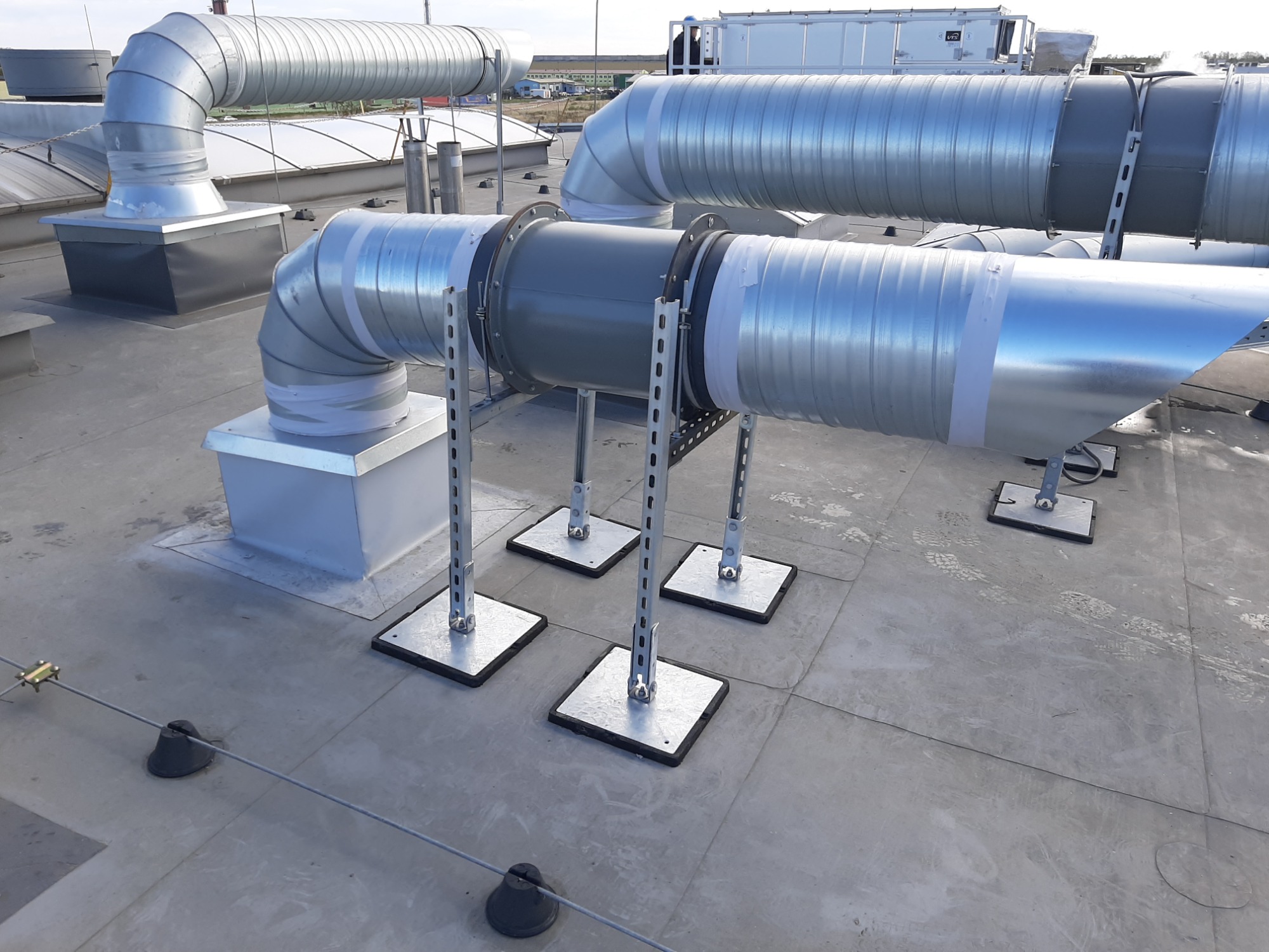 System rur wentylacyjnych ze srebrnej blachy, podtrzymywanych przez metalowe wsporniki na płaskim dachu, z widocznymi śladami stóp na powierzchni dachu.