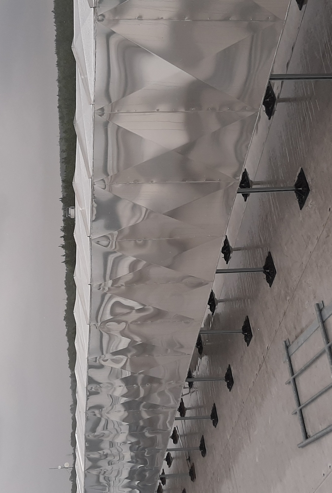 Srebrny kanał wentylacyjny o romboidalnej strukturze, zamontowany na dachu budynku, widoczny w pochmurny dzień z elementami krajobrazu w tle.