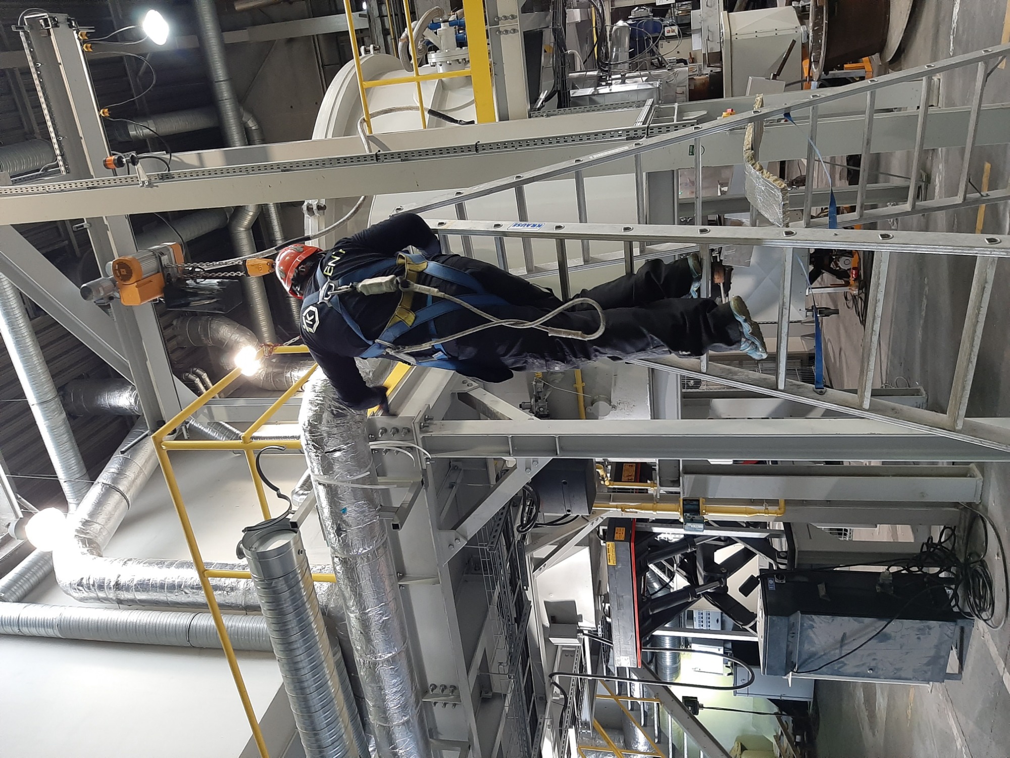 Pracownik w uprzęży na drabinie, instalacja przemysłowa z rurami wentylacyjnymi w tle.