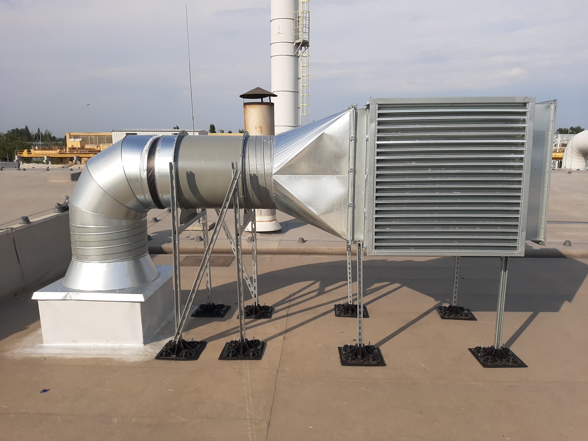 System wentylacyjny z metalowymi kanałami i kratką wentylacyjną na płaskim dachu, widoczne podpory z czarnymi podstawami i elementy mocujące.